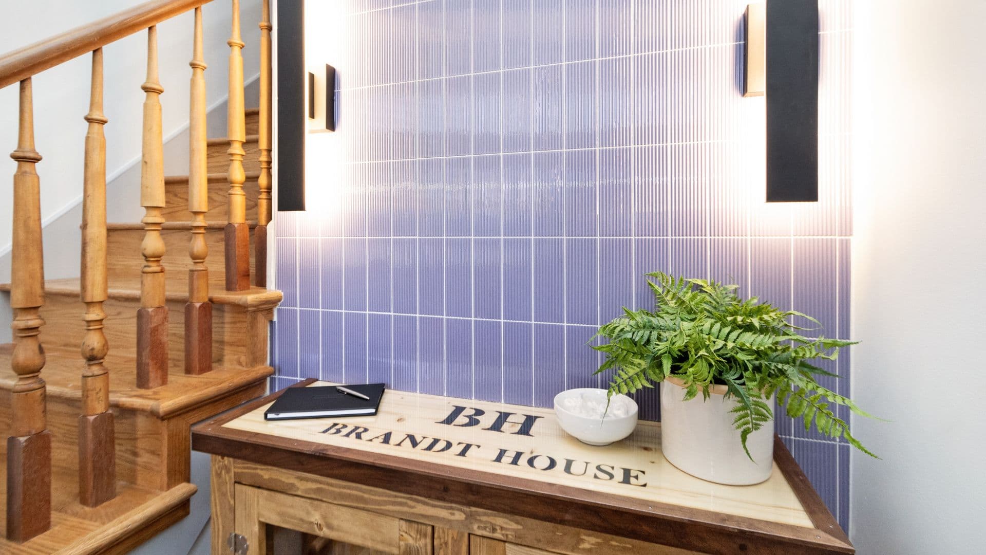 A wooden console table with "BH Brandt House" engraved on it sits against a purple tiled wall, illuminated by wall sconces, with a potted plant and notebook on top.