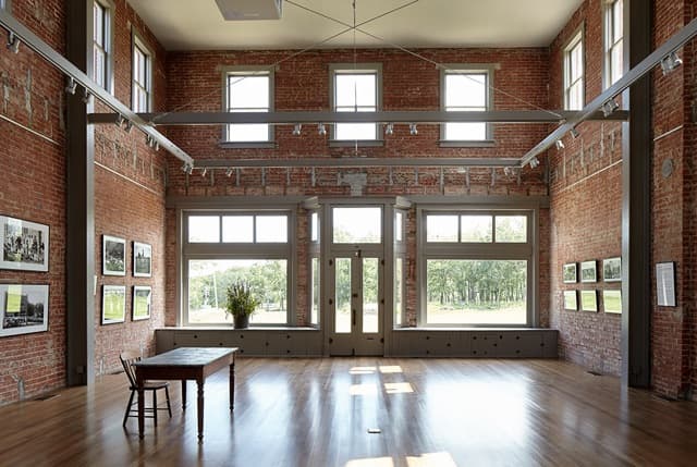 A spacious, well-lit gallery with exposed brick walls and large windows, featuring a central table and framed photographs along the walls.